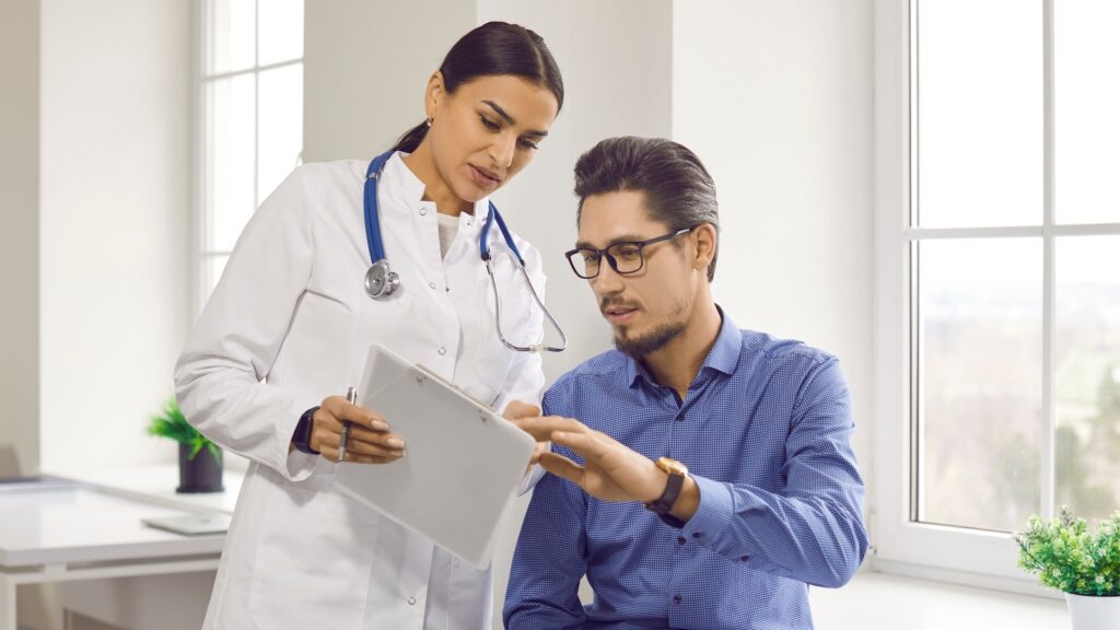 A doctor showing a patient genetic test results on a tablet, with stylized DNA helix‑and‑cancer cell graphics in the background.